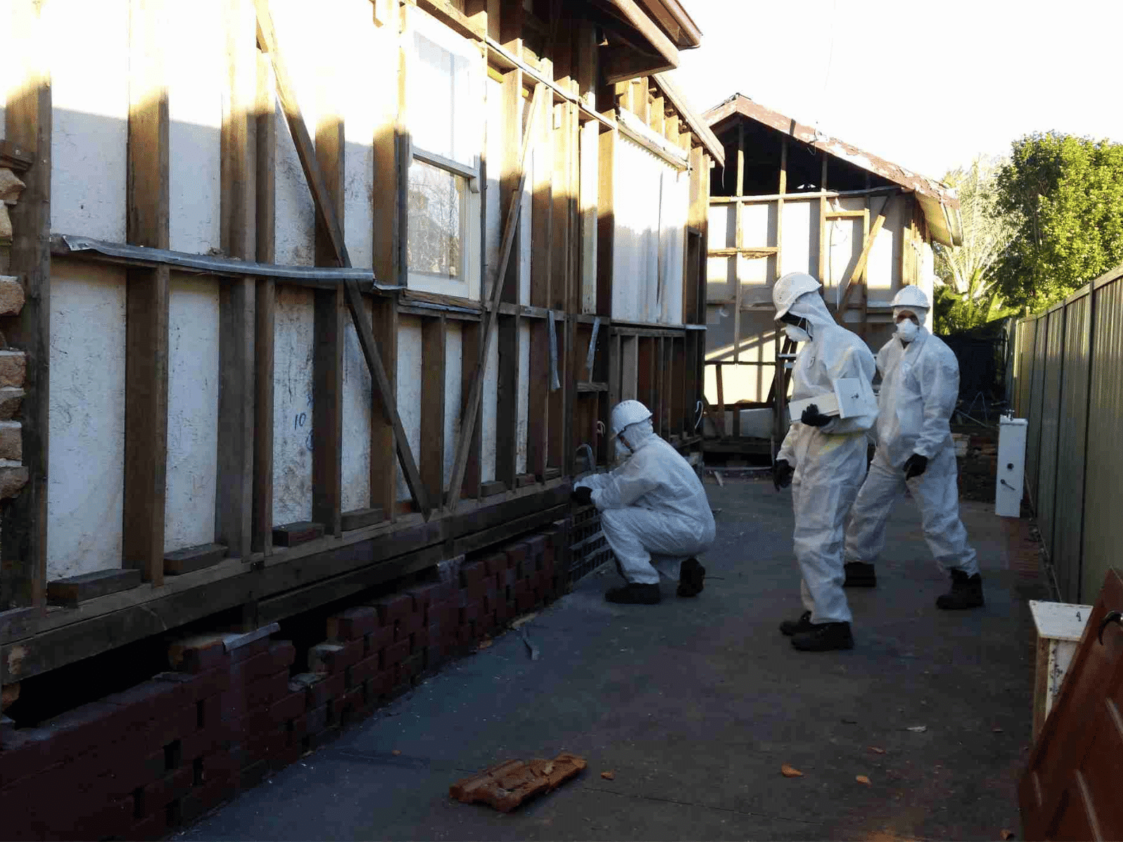 Three people wearing white hazmat suits and masks work on the exterior foundation of a house, inspecting or removing materials from the brick and wooden structure in an outdoor setting.