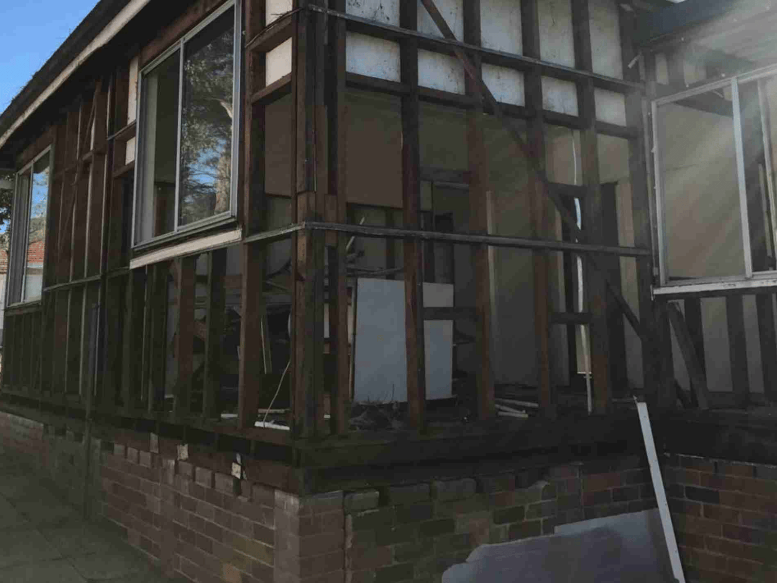 A partially demolished house with exposed wooden framing and missing walls, revealing the interior. Some windows remain intact, while debris is visible inside. Sunlight shines onto the structure.