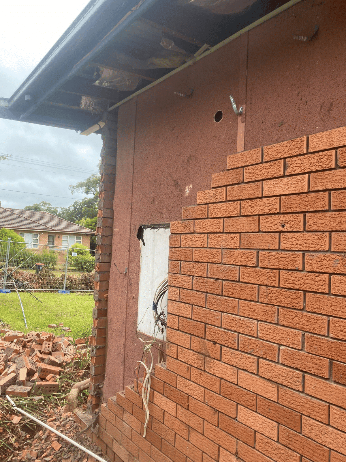 A partially demolished brick wall reveals electrical wires and a panel on the outer side of a house; bricks and debris are scattered on the ground, with a lawn and neighboring house in the background.