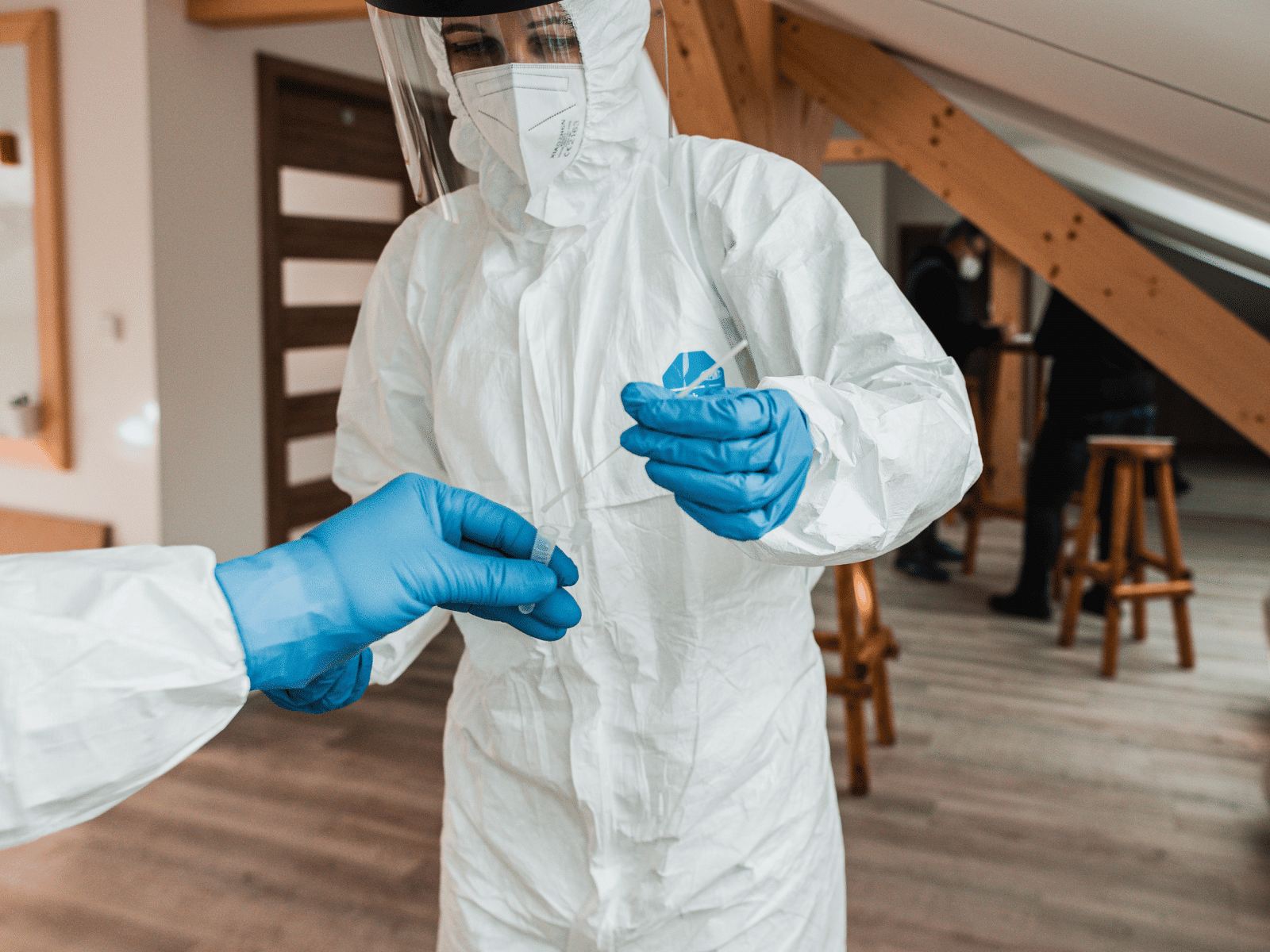 Two people wearing full protective suits, gloves, and face shields handle a swab sample in a bright indoor space with wooden beams and stools in the background.