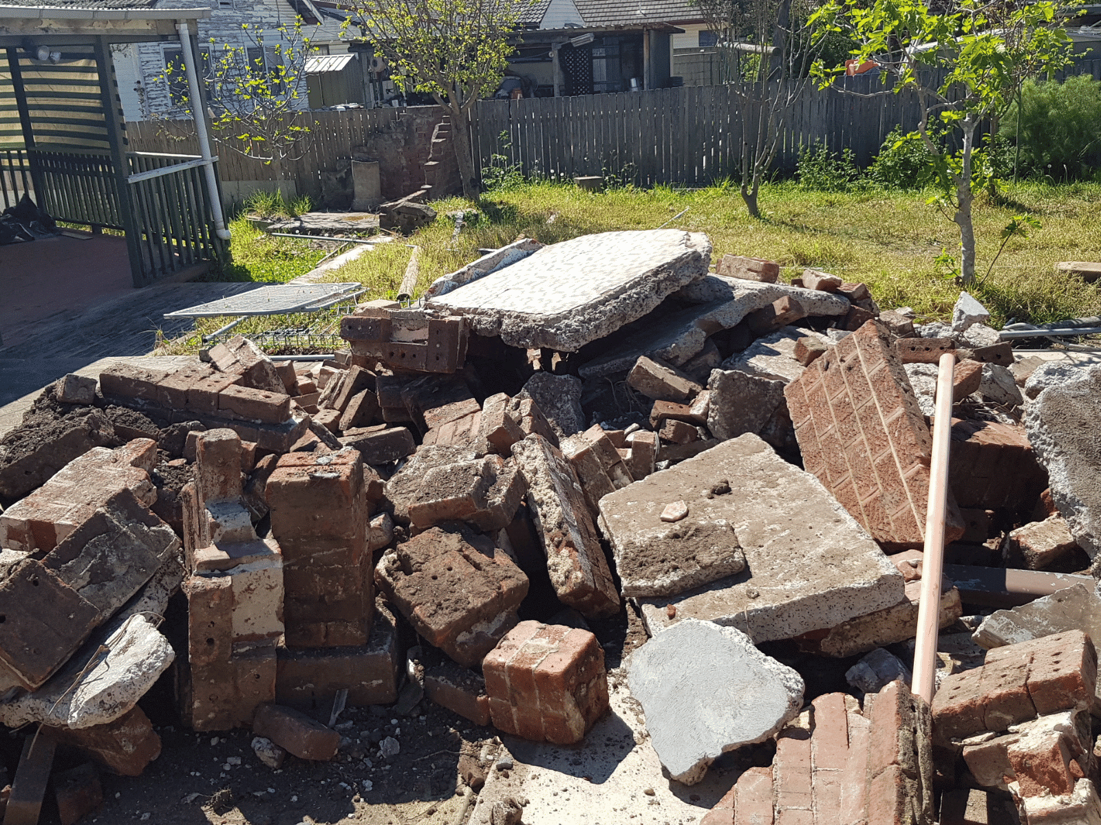 A pile of broken bricks, concrete, and rubble sits in a yard with grass, trees, and a wooden fence in the background. Sunlight casts shadows across the debris.