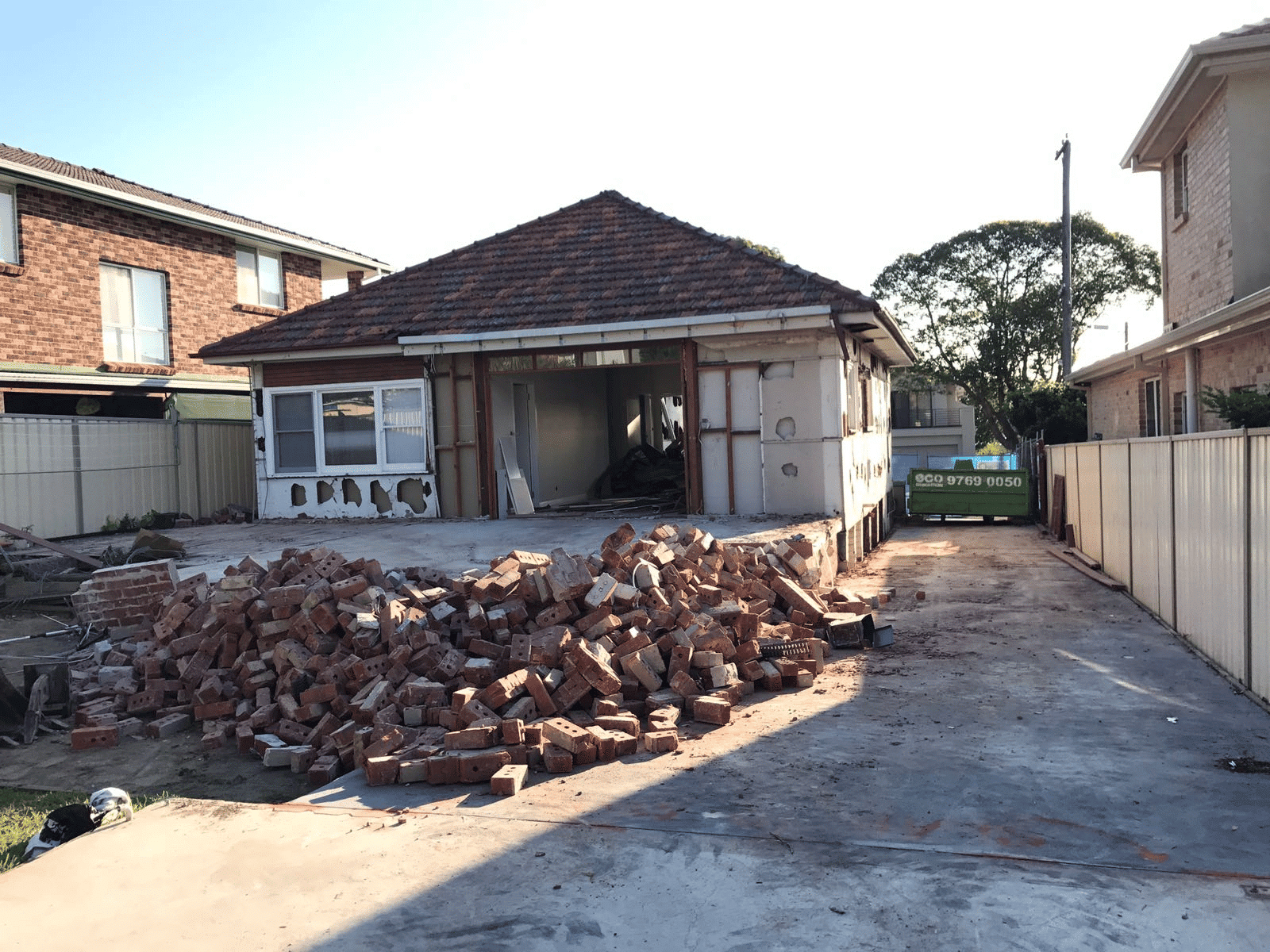 A worn, empty room with peeling blue walls, exposed ceiling beams, and a wooden floor. Cleaning tools like a broom and mop are in one corner, and sunlight comes through two windows and a glass door.