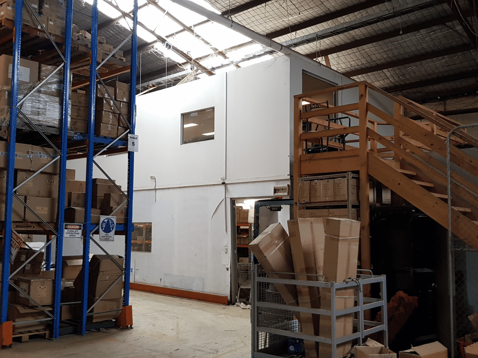 A warehouse interior with stacked shelves of boxes, a white office structure with windows, and a wooden staircase leading to a second floor. Cardboard boxes are placed in carts and on the floor nearby.