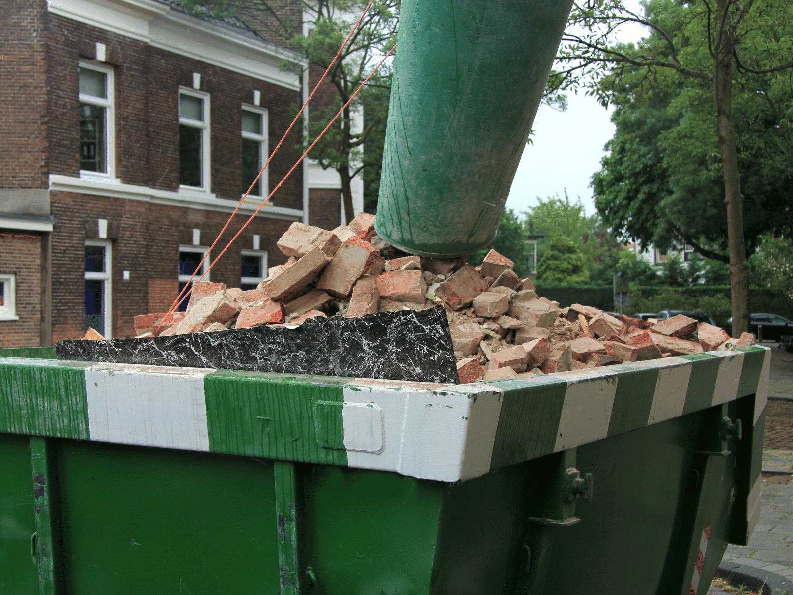 A green dumpster filled with broken bricks and debris sits outdoors near a brick building and trees, with a green chute feeding more rubble into the container.
