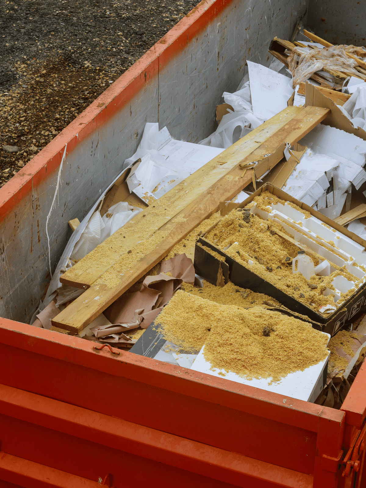 A red dumpster filled with construction debris, including wooden planks, white papers, cardboard, and scattered sawdust inside a plastic container.