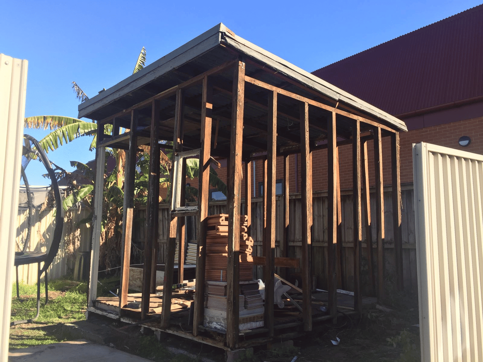 A wooden shed frame stands without walls, revealing stacks of construction materials inside. The structure sits in a fenced yard with grass, a banana plant, and brick buildings in the background under a clear blue sky.