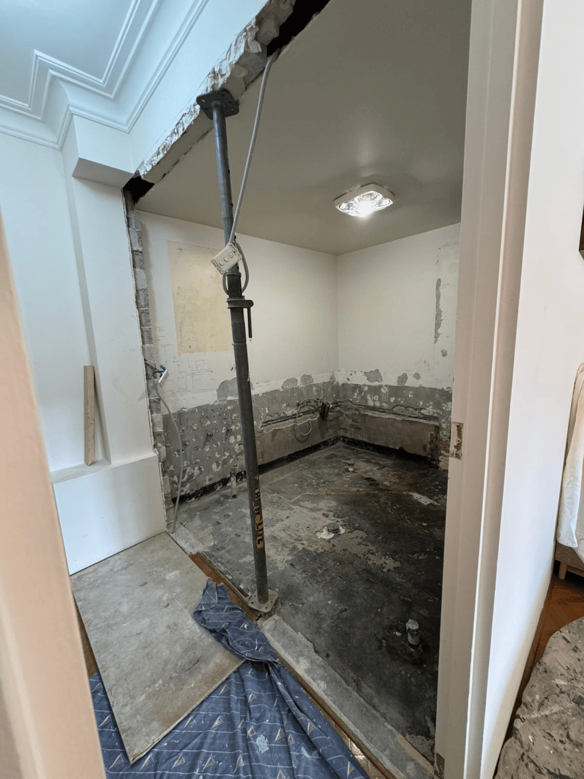 A partially demolished room with exposed walls and floor, construction support pole, and debris. The ceiling light is on, and a blue protective mat is laid at the entrance.