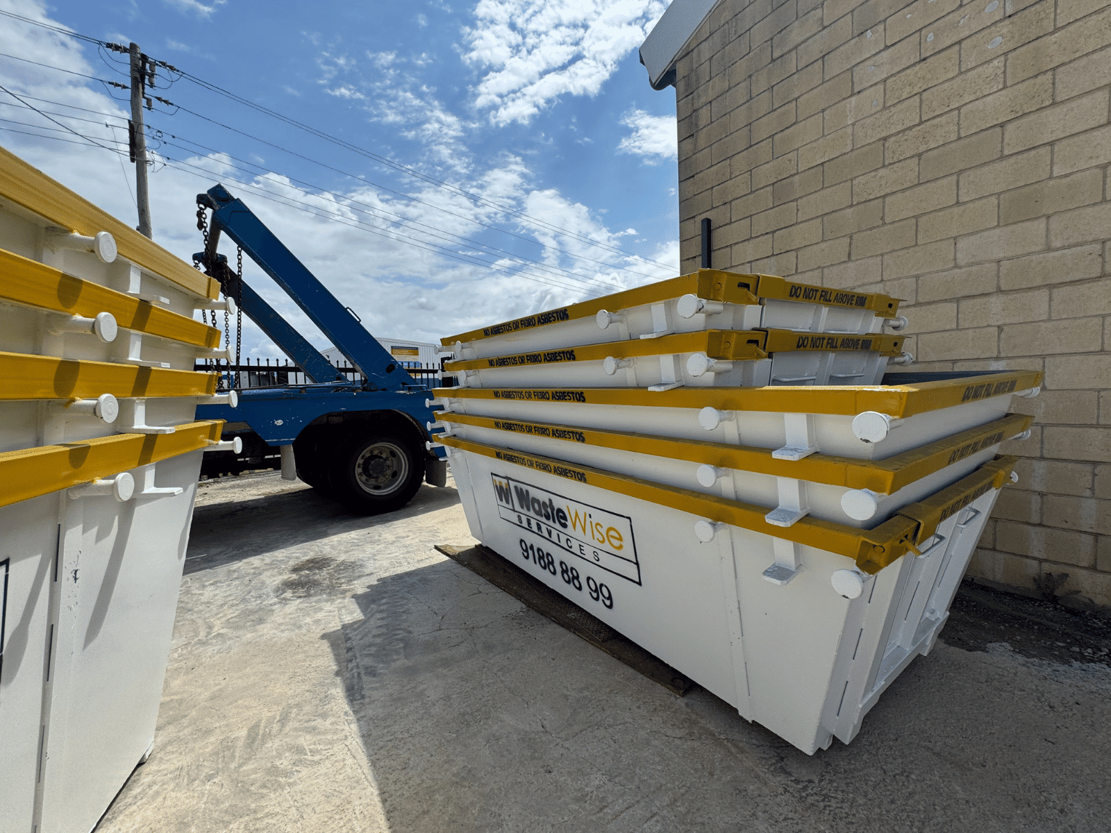 A white skip bin with yellow trim sits on the grass in front of a single-story brick house with a covered porch. A driveway and parked cars are visible beside the house on a sunny day.