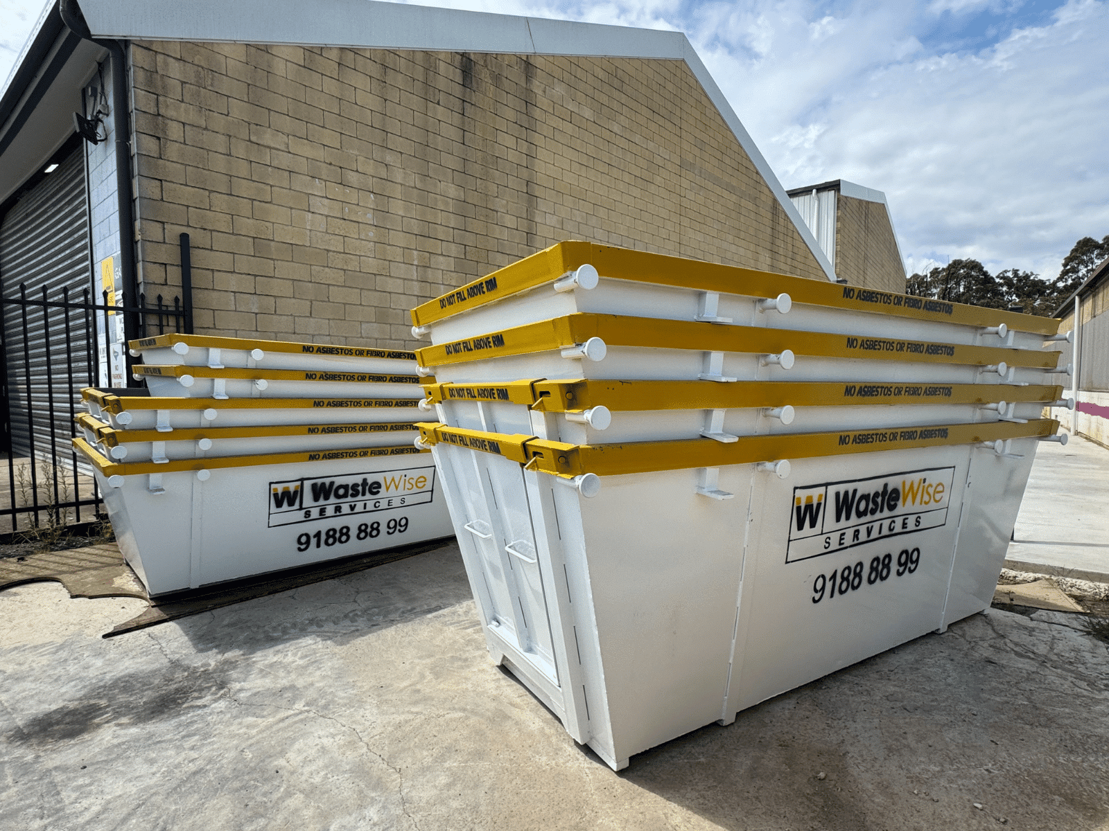 Several white and yellow WasteWise Services dumpsters are stacked outside a building with a beige brick wall and metal shutter. The company logo and phone number are visible on the bins.