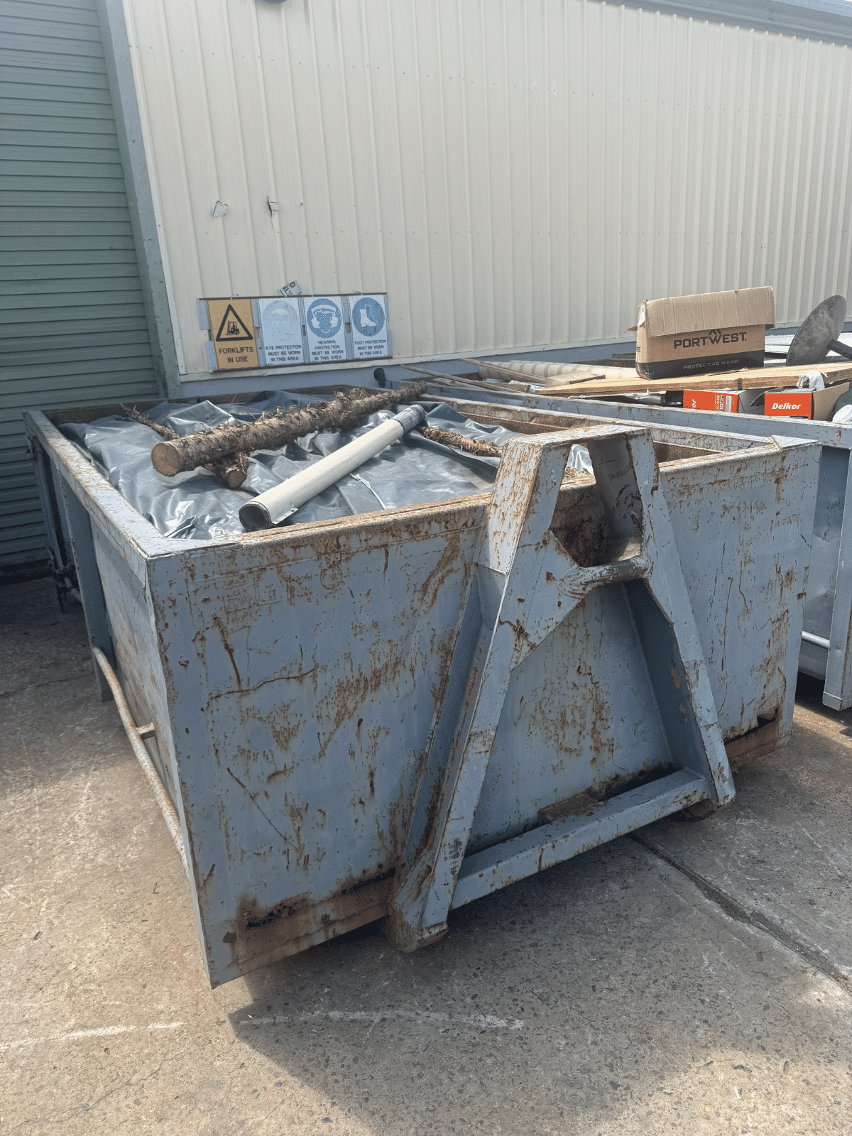 A large, rusty metal dumpster filled with pipes and other debris sits outside a corrugated metal building, with warning signs and cardboard boxes nearby on a concrete surface.