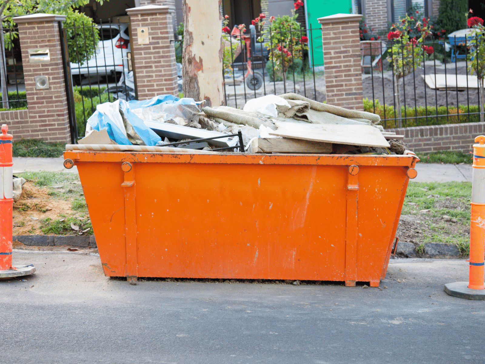 An orange dumpster filled with construction debris and trash is placed on a street between two orange traffic cones, with a brick fence and greenery in the background.