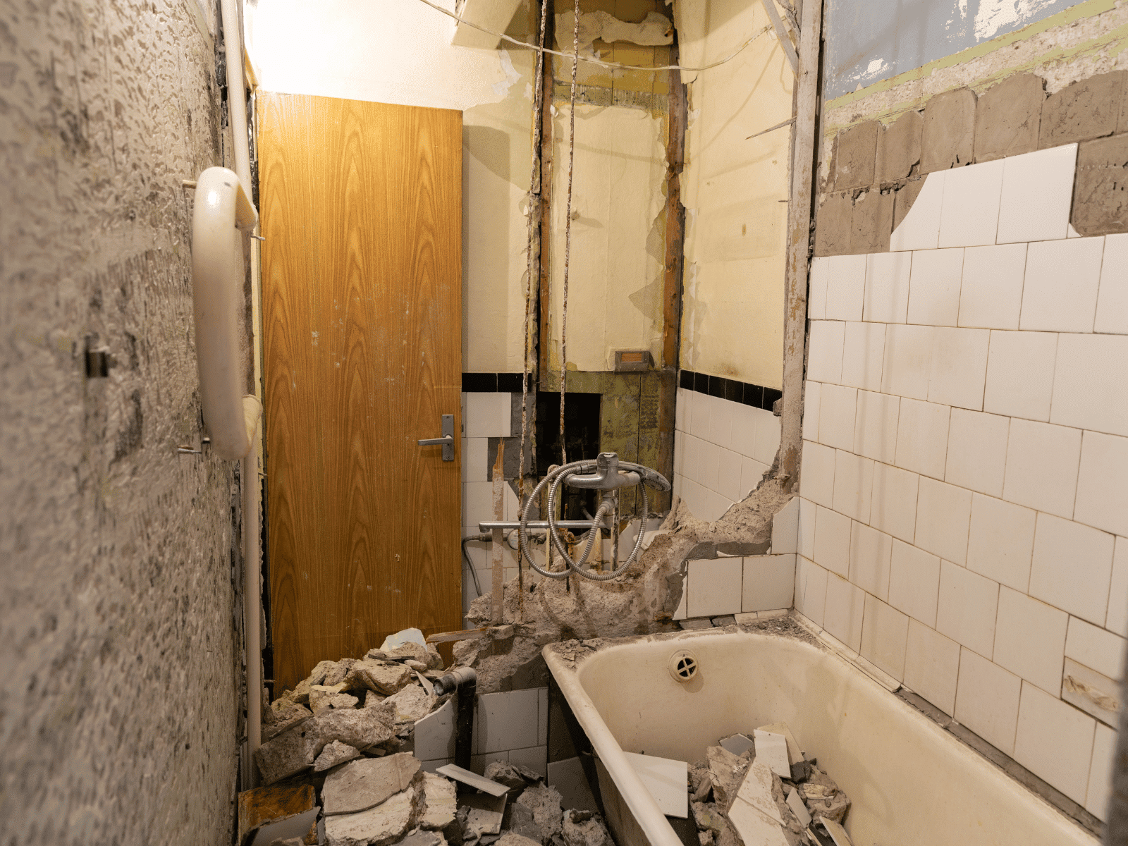 A bathroom under renovation with broken tiles, rubble on the floor, exposed pipes, and a partially demolished wall above a dirty bathtub. The wooden door is closed, and the walls show signs of damage and stripped plaster.