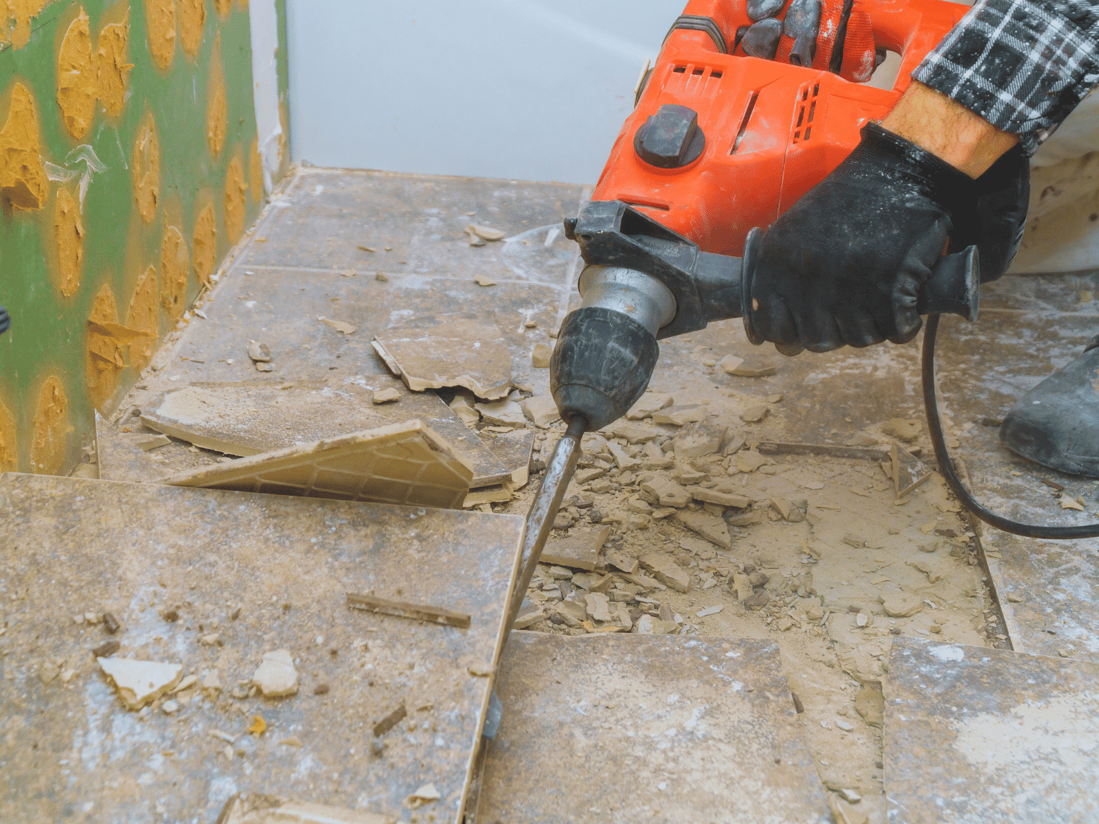 A person wearing black gloves uses a red power drill with a chisel attachment to remove broken tiles from a floor, with debris and dust scattered around the work area.