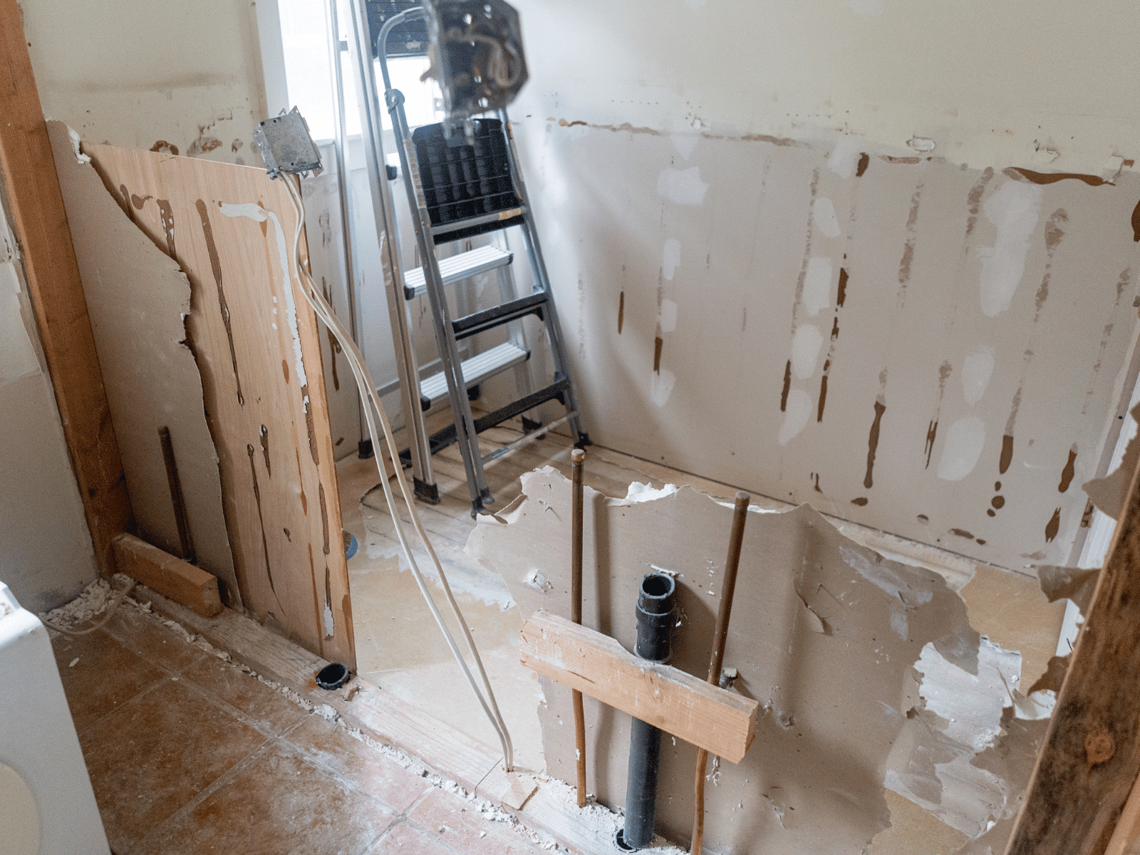 A room under renovation with exposed wall studs, plumbing pipes, and electrical wiring. Drywall is partially removed, and a ladder stands in the background. Construction materials and tools are scattered on the floor.