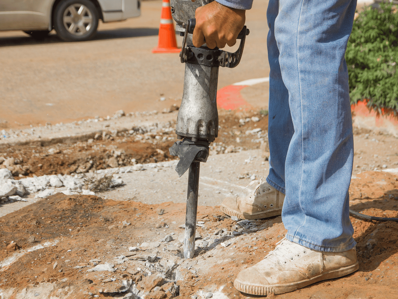 A person in jeans and sneakers uses a jackhammer to break up concrete on a sidewalk, with debris scattered around and a traffic cone visible in the background.