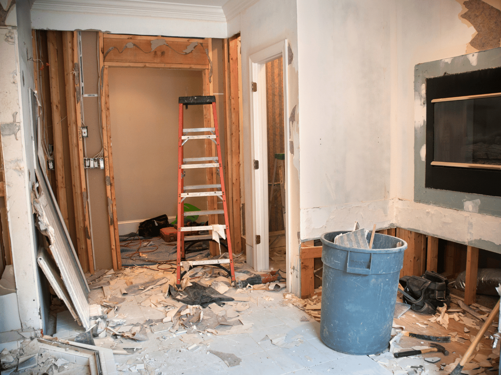 A room undergoing renovation with exposed wall studs, a red ladder, debris on the floor, a large trash bin, and unfinished drywall. Tools and electrical wiring are visible in the background.